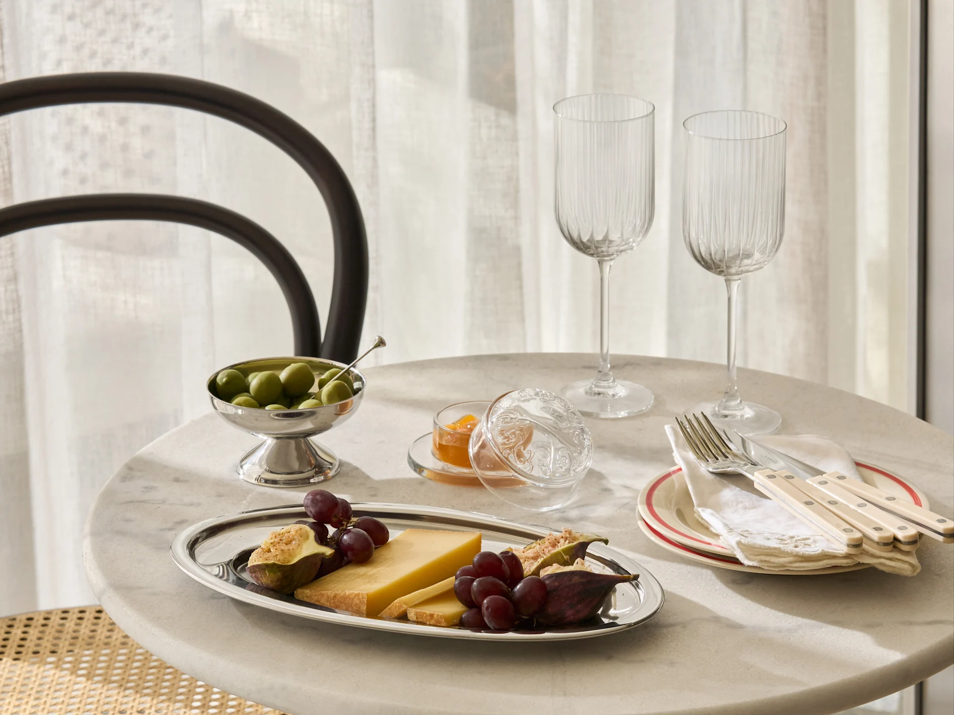 Ein Marmortisch mit einer Silberplatte Käse, Trauben, Feigen, einer Schale Oliven, Marmelade, zwei gerippten Gläsern und Besteck auf Tellern.
