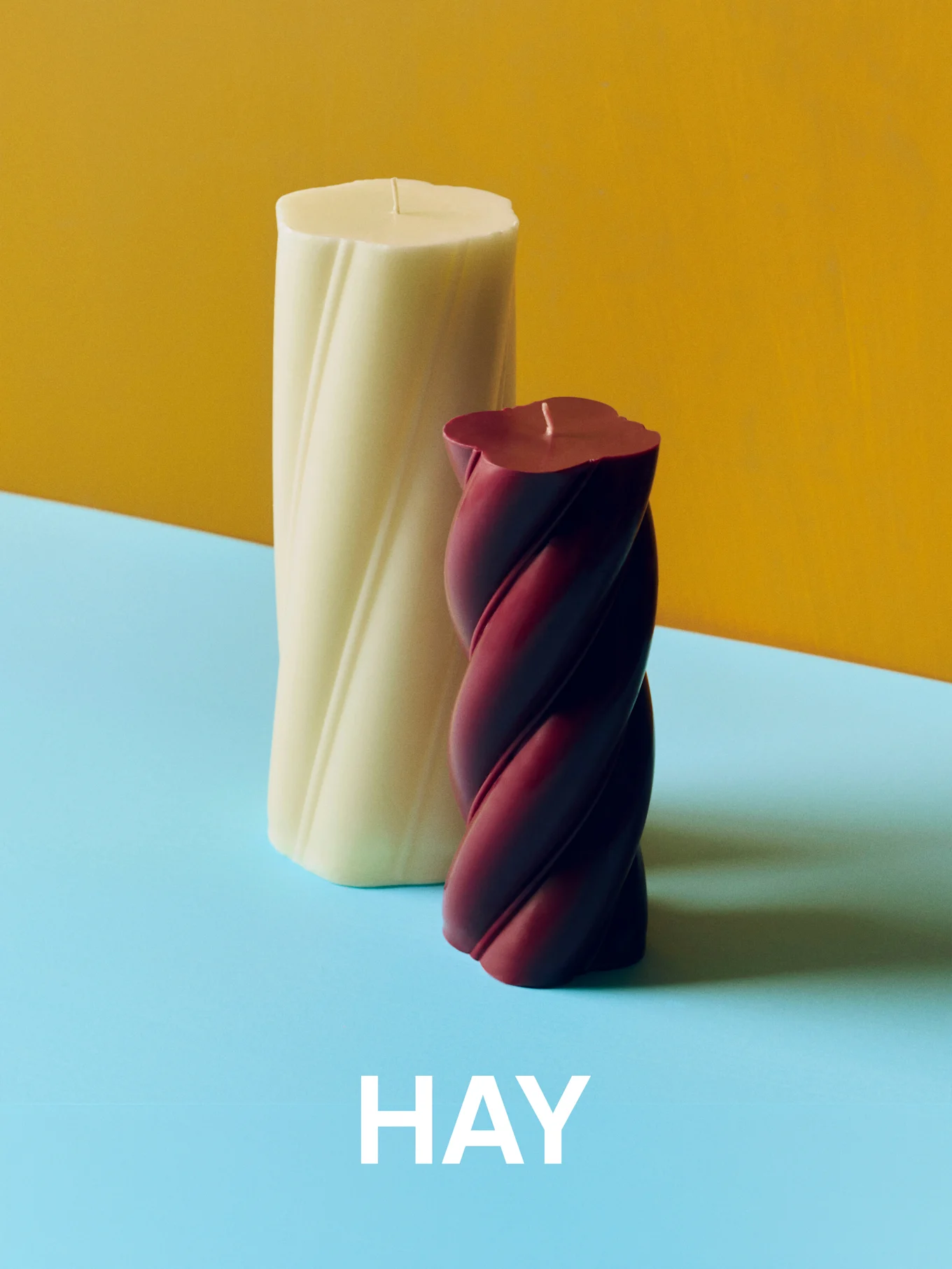 Cremefarbene und burgunderfarbene gedrehte Kerzen auf gelbem und blauem Hintergrund mit dem Text 'HAY'.