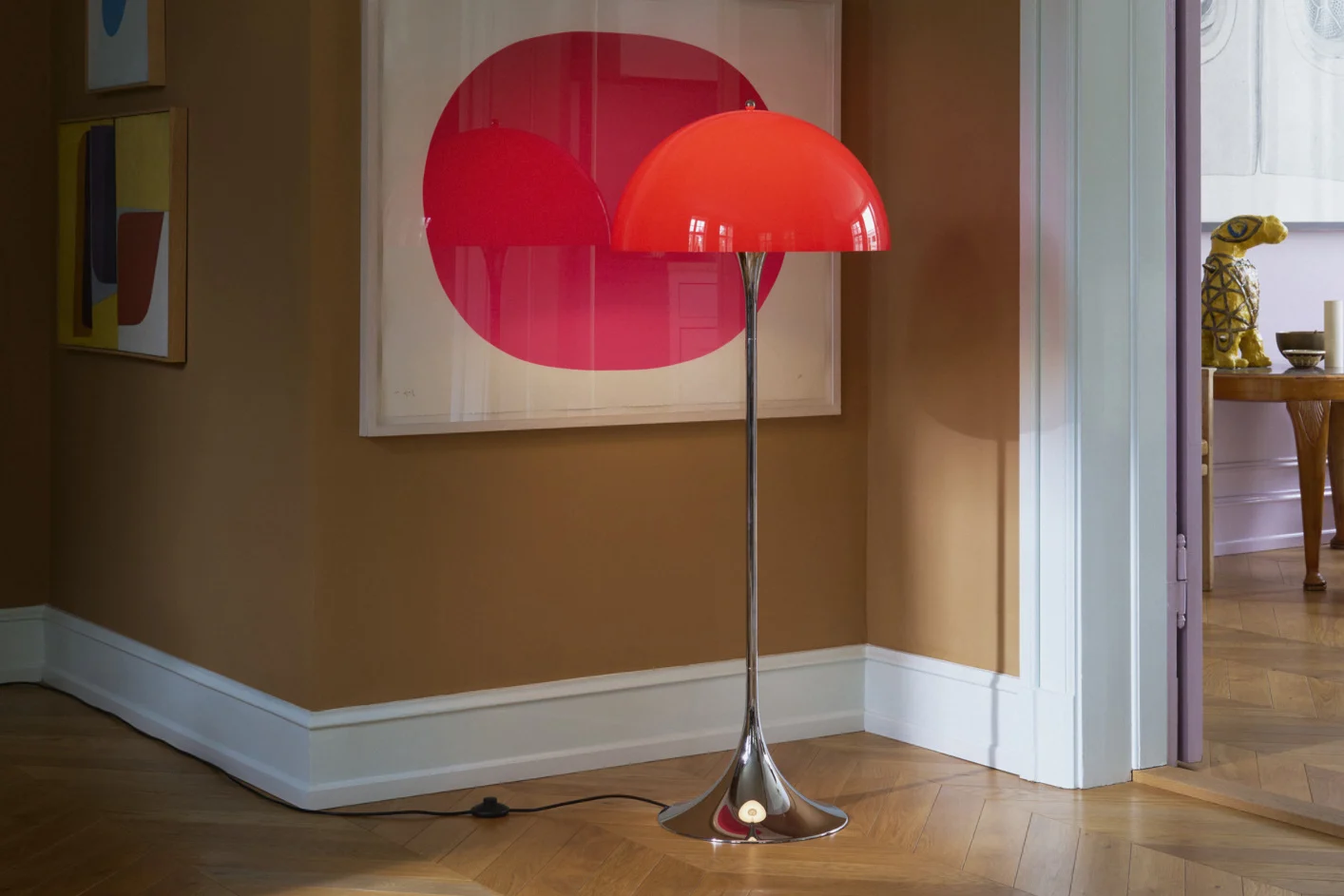 Eine leuchtend rote gewölbte Stehlampe mit Chromständer steht auf einem Holzfußboden in einem Raum mit braunen Wänden und moderner Kunst.