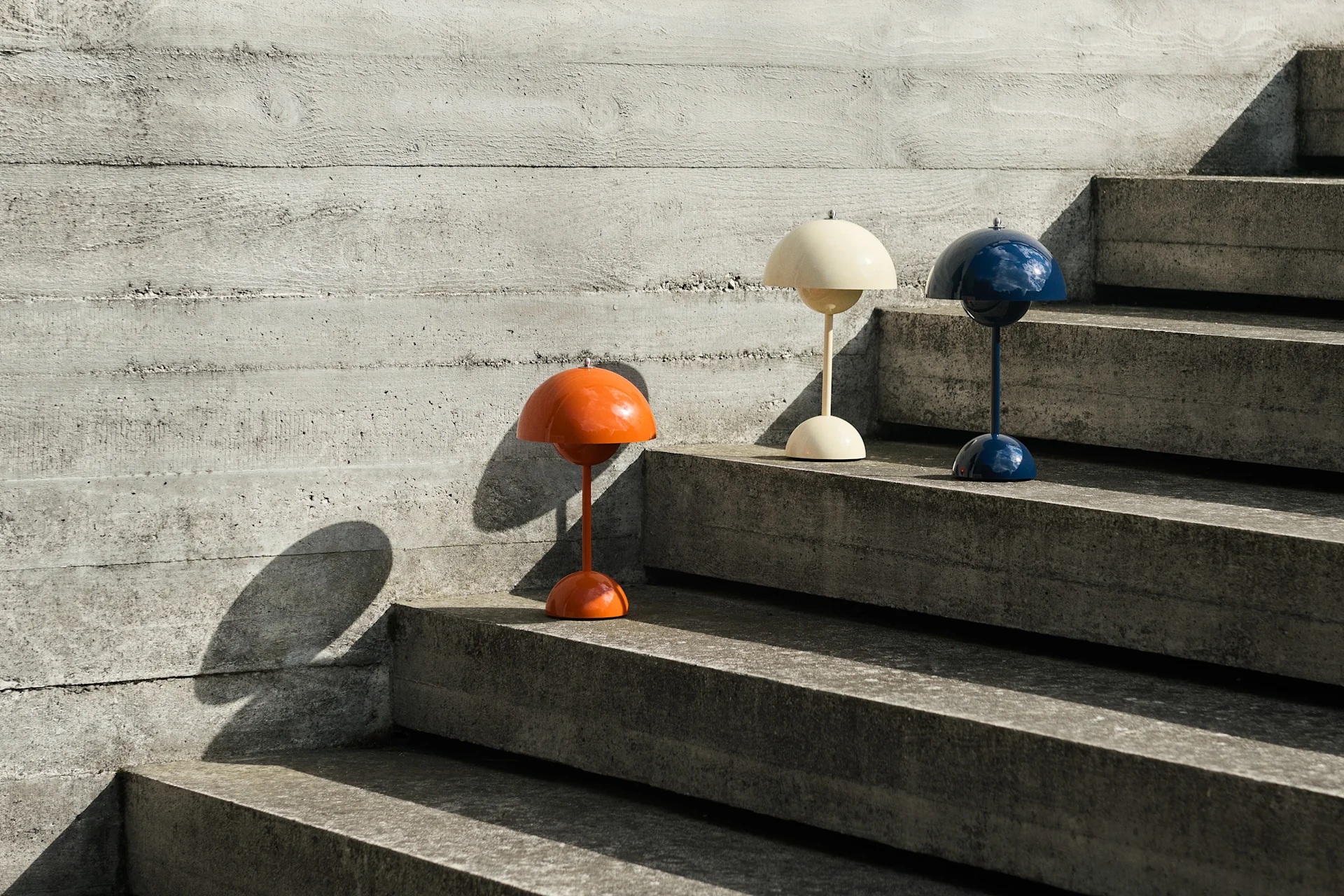 Drei kuppelförmige Tischlampen in Orange, Creme und Blau auf Betontreppen neben einer Betonwand, die Schatten werfen.