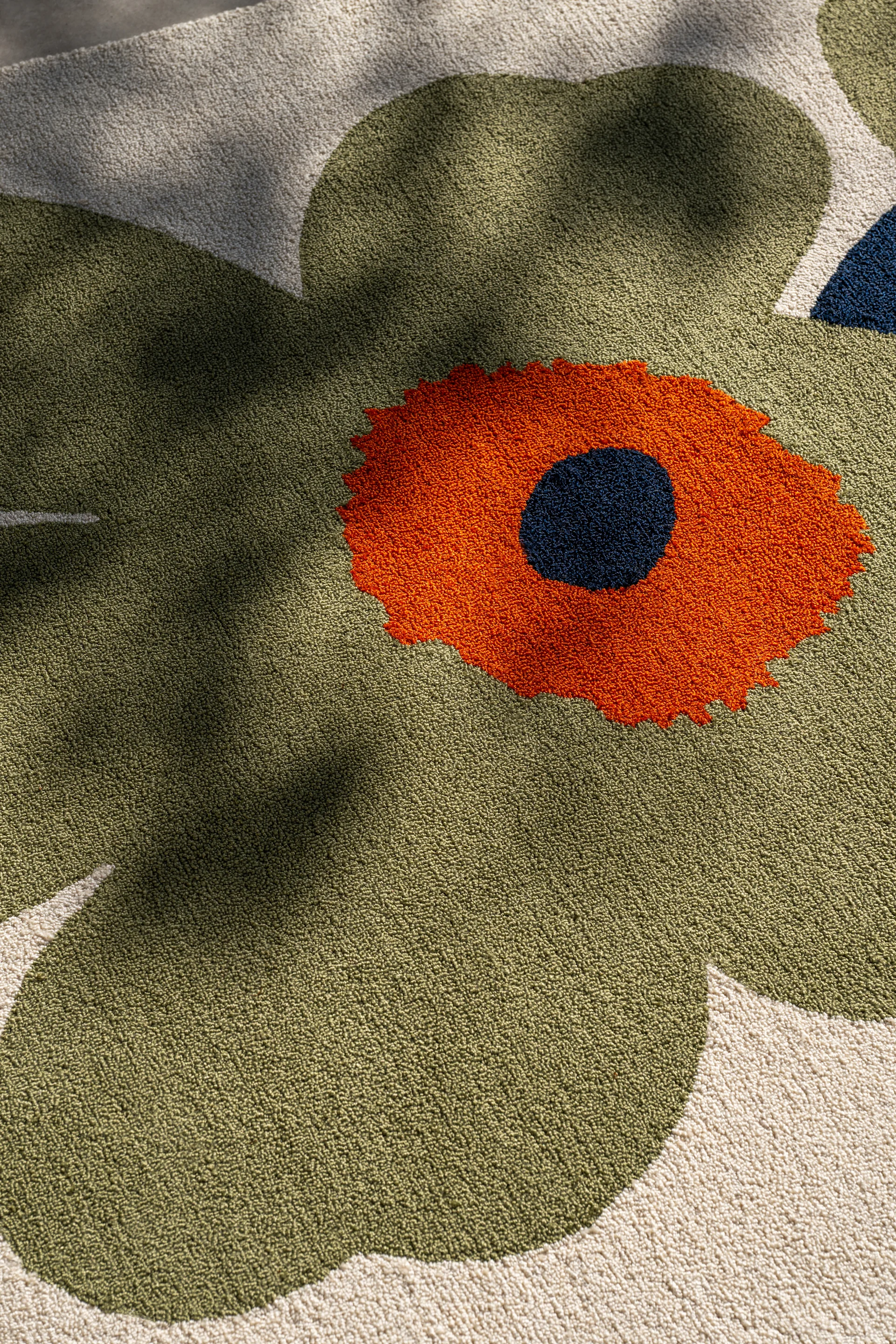 Nahaufnahme eines Teppichs mit großem, stilisiertem Blumenmuster. Olivgrüne Blütenblätter, orange Mitte und ein dunkelblauer Punkt auf cremefarbenem Hintergrund.
