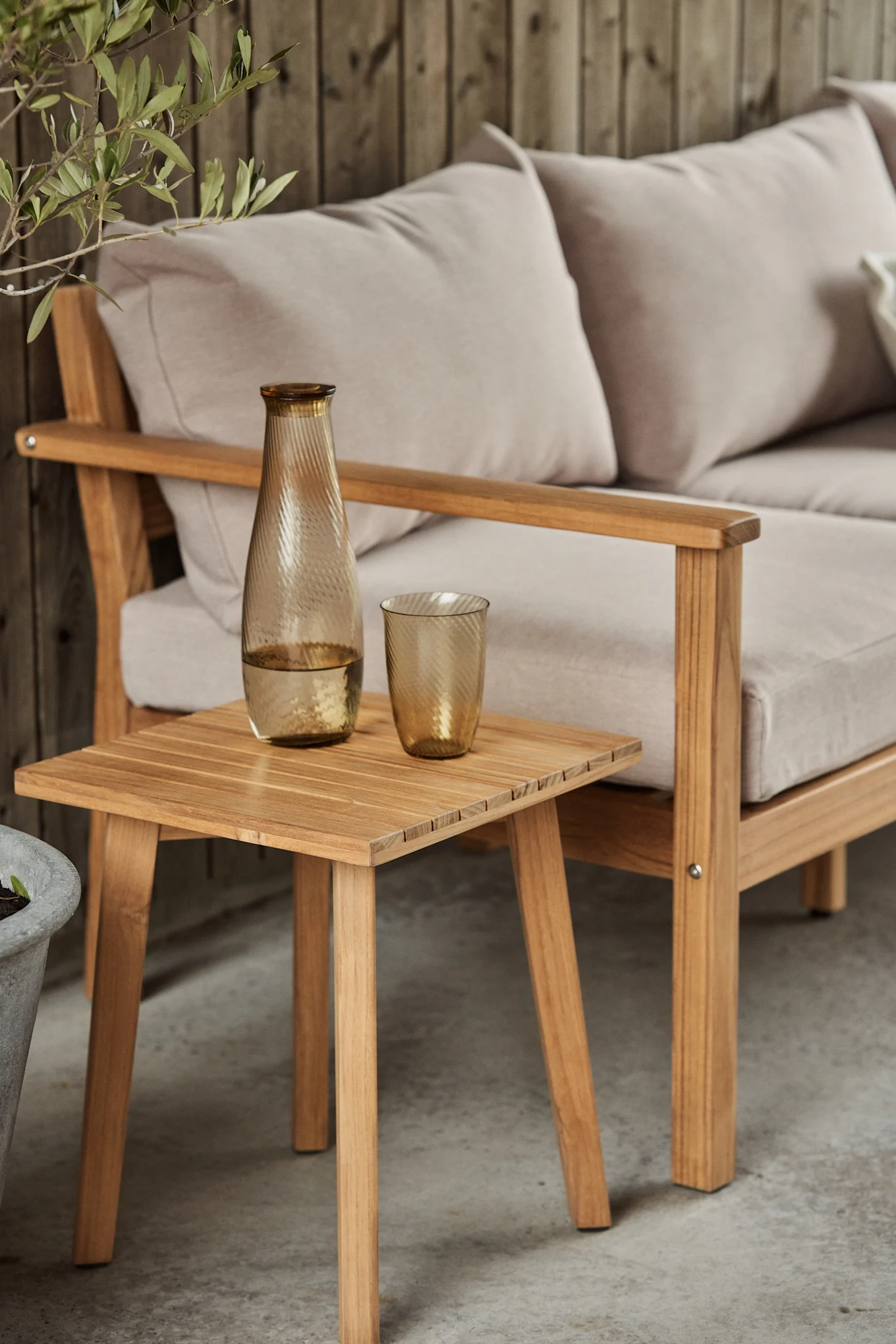 Holz-Gartensofa mit hellen Kissen, ein Beistelltisch aus Holz mit bernsteinfarbener geriffelter Glaskaraffe und passendem Glas.