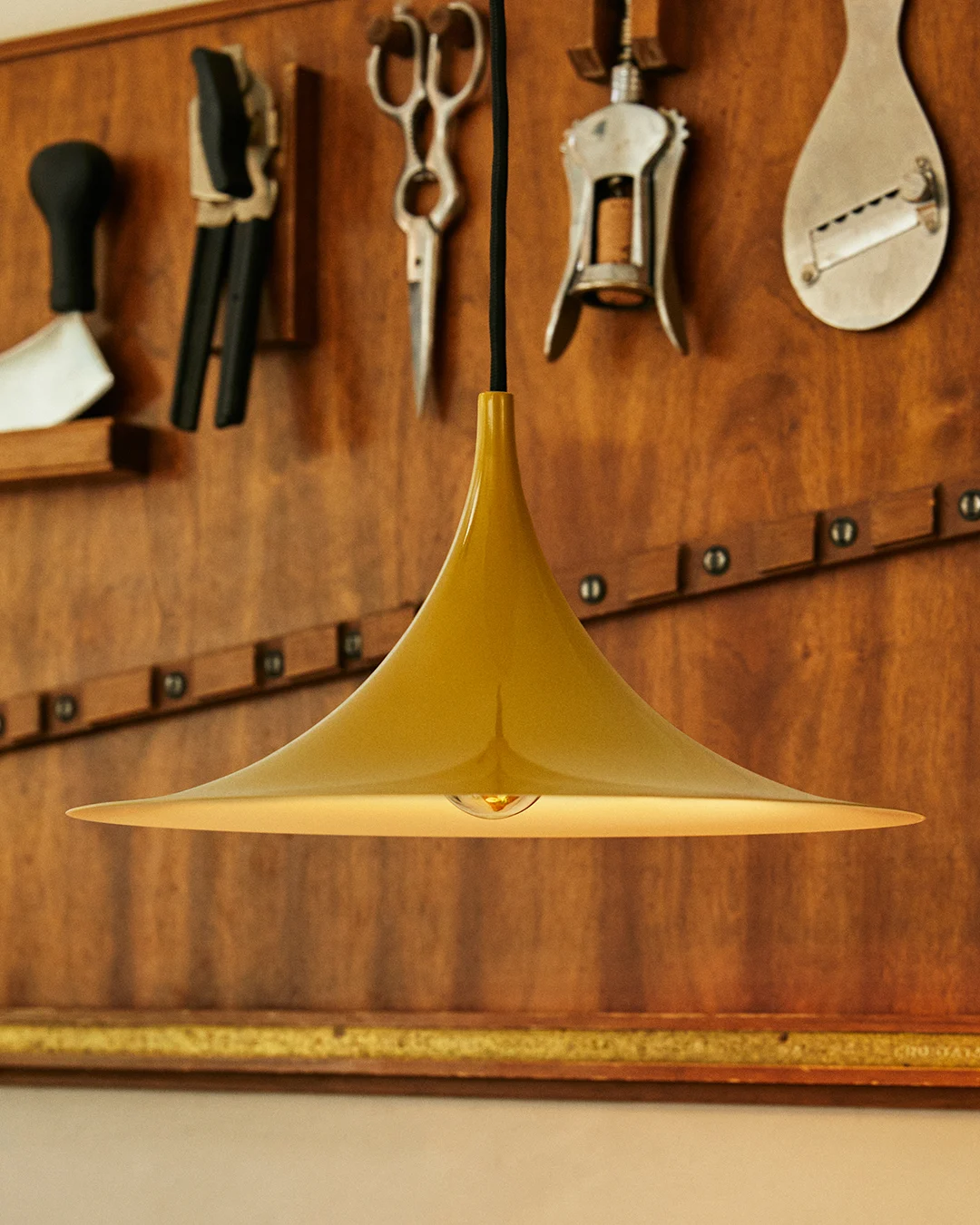 Goldene glockenförmige Pendelleuchte mit sichtbarer Glühbirne, vor einer Holzwand mit Werkzeugen.