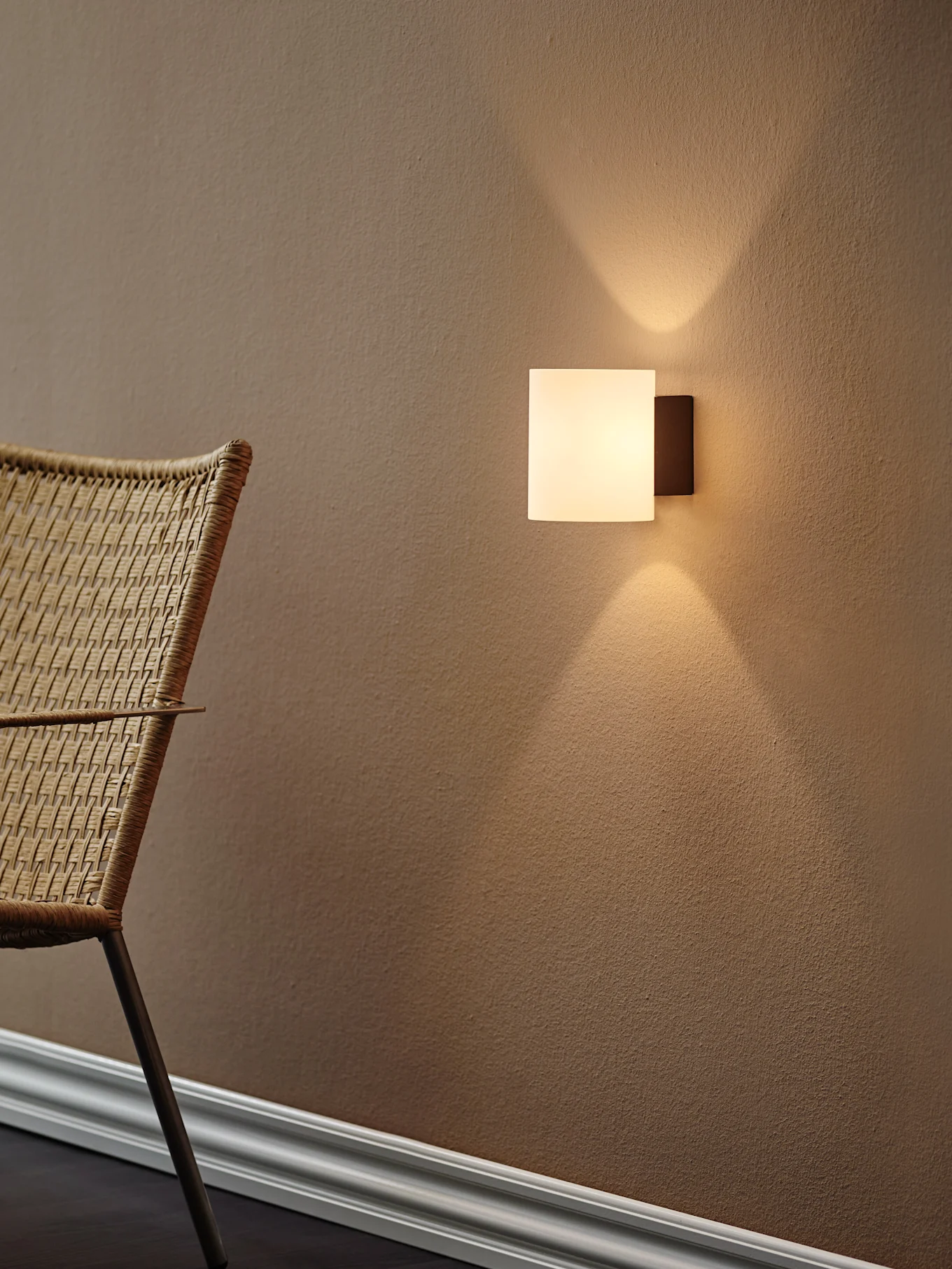 Moderne quadratische Wandleuchte, die warmes Licht nach oben und unten auf eine strukturierte braune Wand wirft, mit einem geflochtenen Stuhl links.