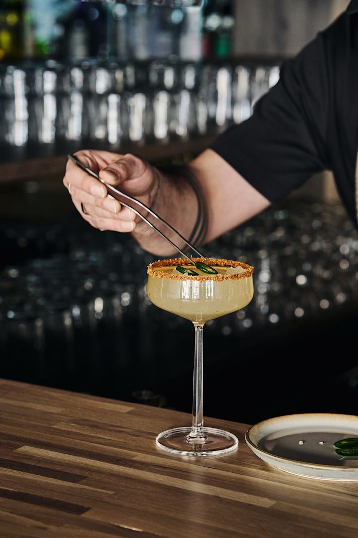 Perfekt geeignet für alkoholfreie Cocktails: Die Essence Cocktailgläser von Iittala mit einem Drink, dekoriertem Rand und mit einer Zange garniert mit Jalapeño-Scheiben.