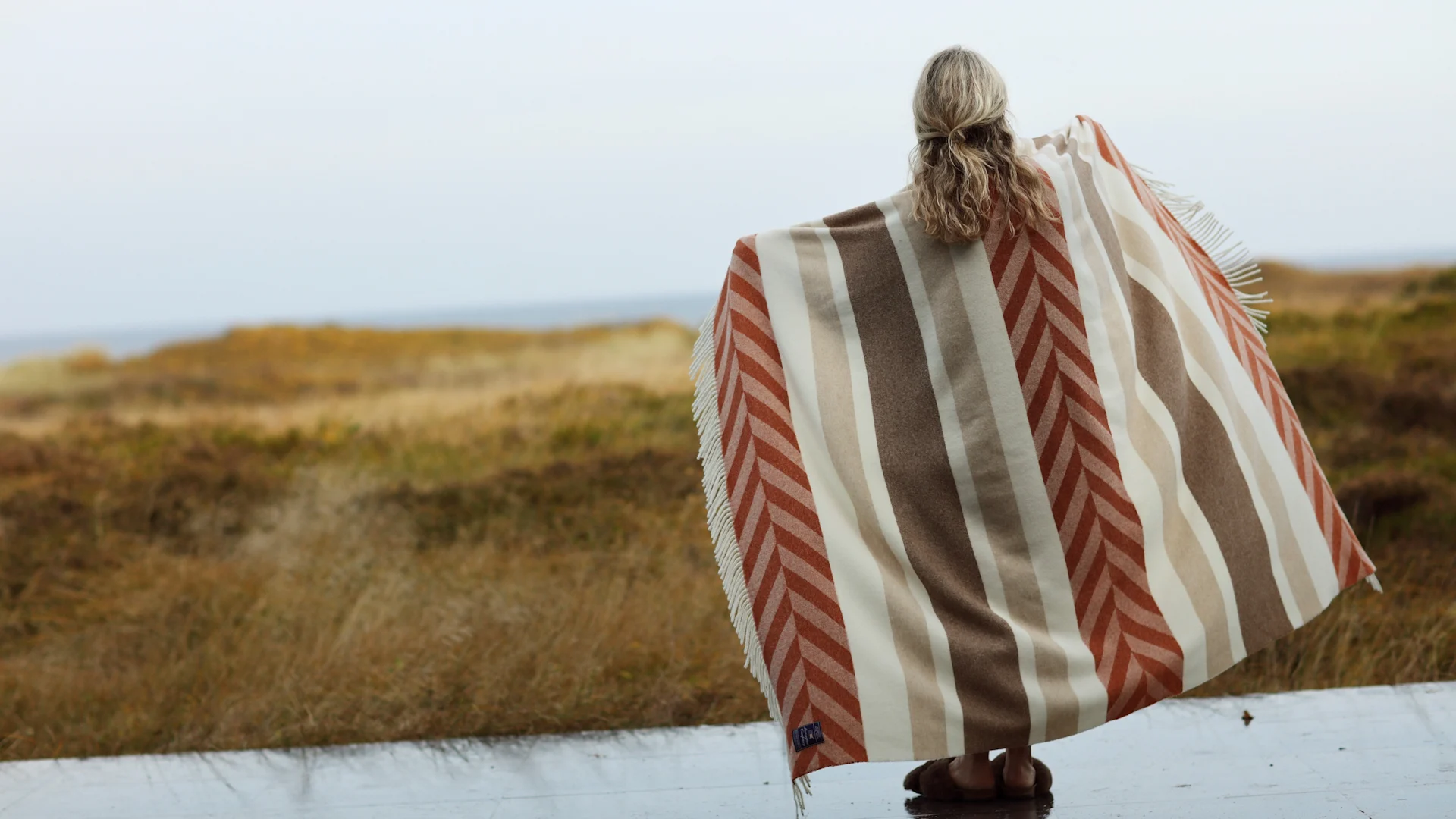 Unverzichtbar für den Herbst: Eine kuschelige Wolldecke. Hier steht eine Frau auf einer Terrasse vor einer weiten Landschaft mit der Herringsbone Striped Wolldecke von Lexington.