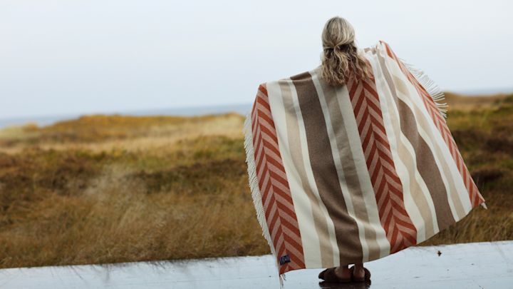 Unverzichtbar für den Herbst: Eine kuschelige Wolldecke. Hier steht eine Frau auf einer Terrasse vor einer weiten Landschaft mit der Herringsbone Striped Wolldecke von Lexington.