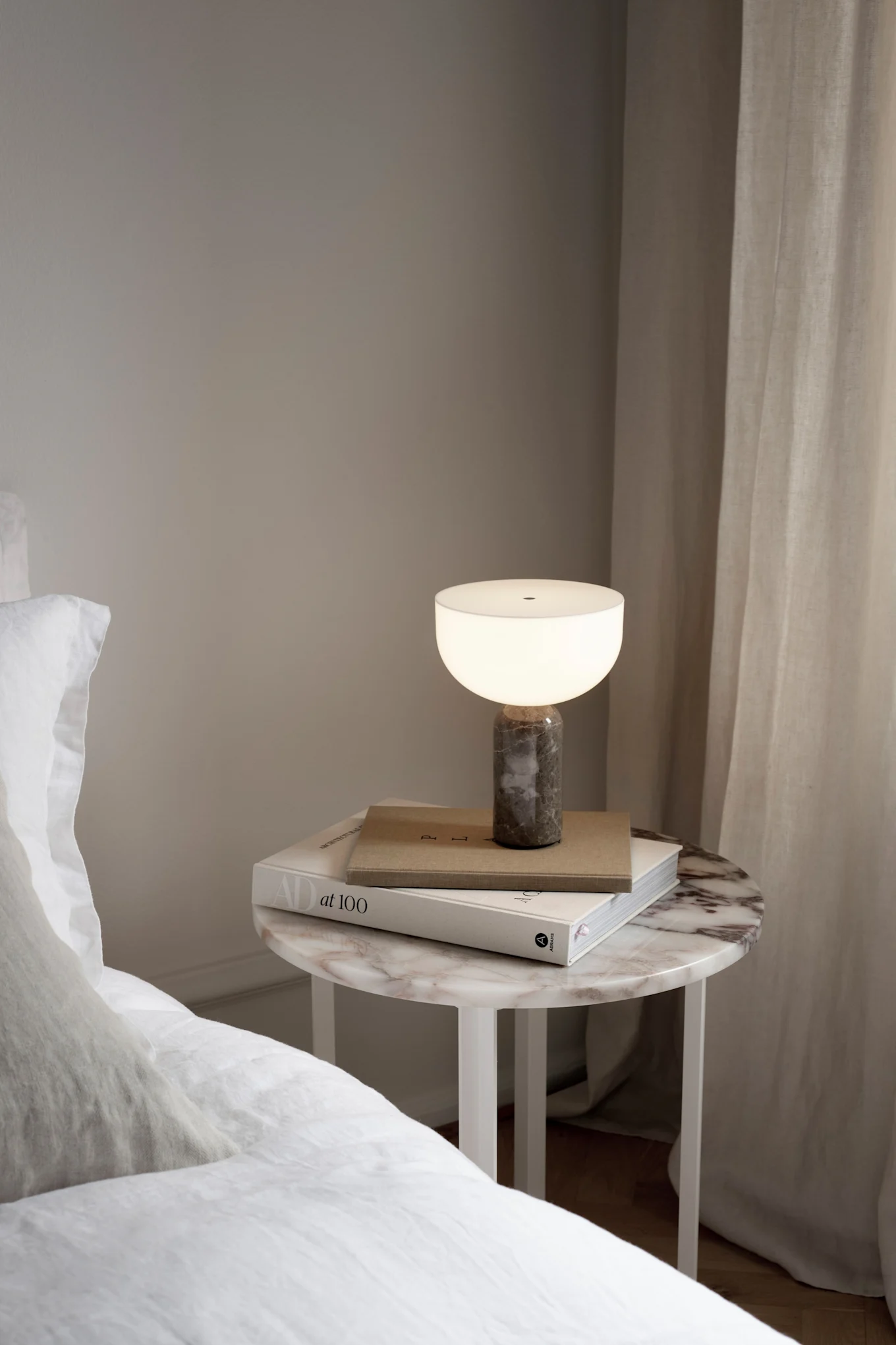 Die Akku-Tischleuchte Kizu von New Works steht auf einem Nachttisch neben einem Bett.