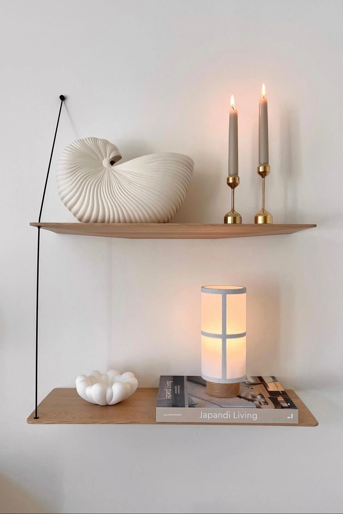 Zwei Holzregale mit einer Nautilusmuschel, brennenden Kerzen, einem Japandi Living Buch, einer gerüschten Schale und einer leuchtenden Lampe.