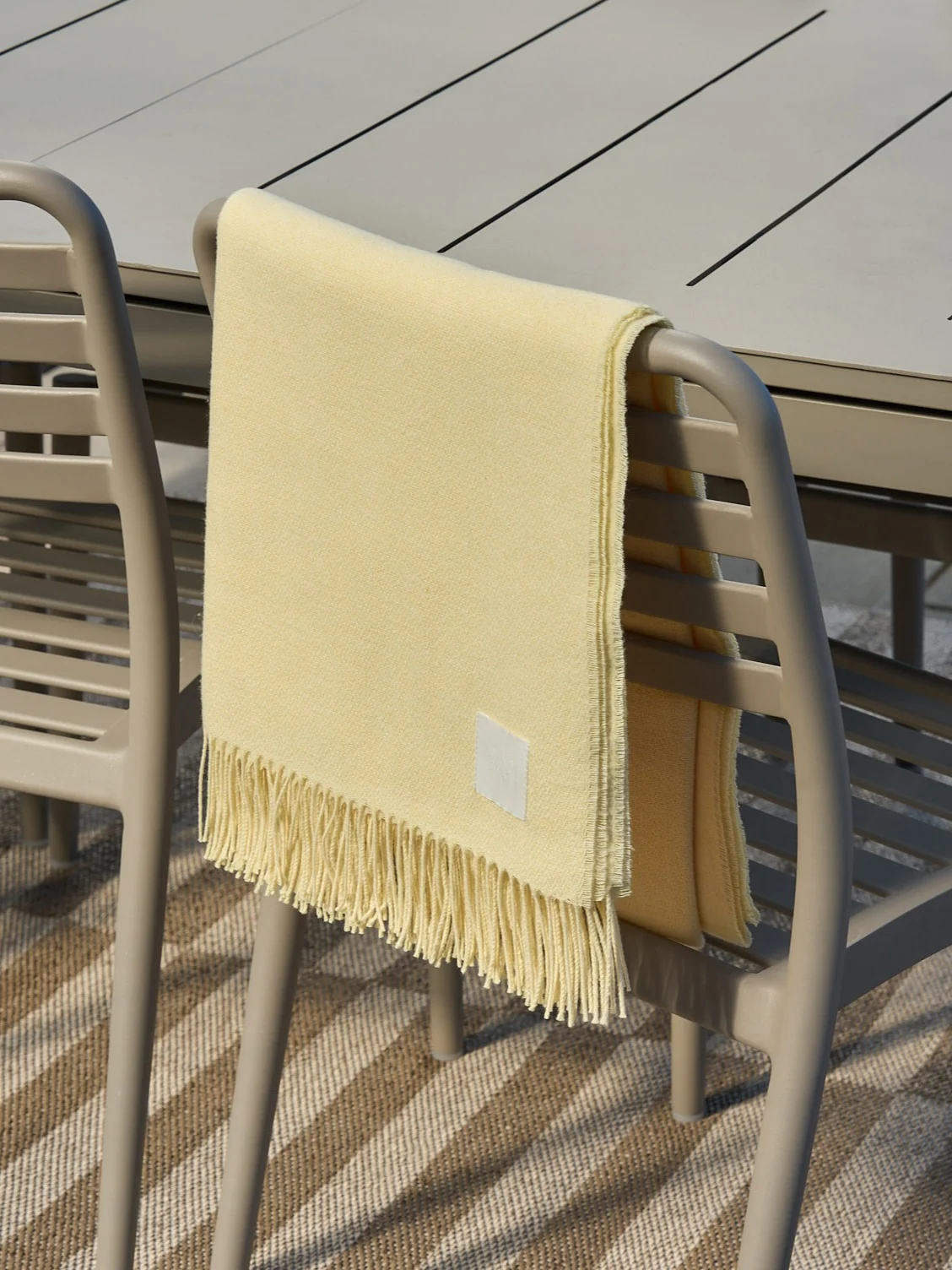 Hellgelbe Decke mit Fransen über einem beigefarbenen Outdoor-Stuhl drapiert.