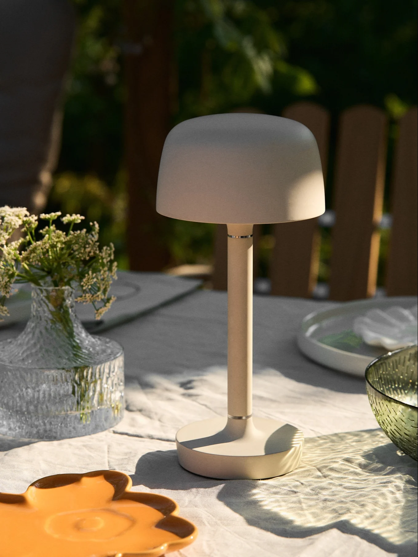 Beige tragbare Tischlampe auf einem Gartentisch mit Blumenvase und gesprenkeltem Sonnenlicht.