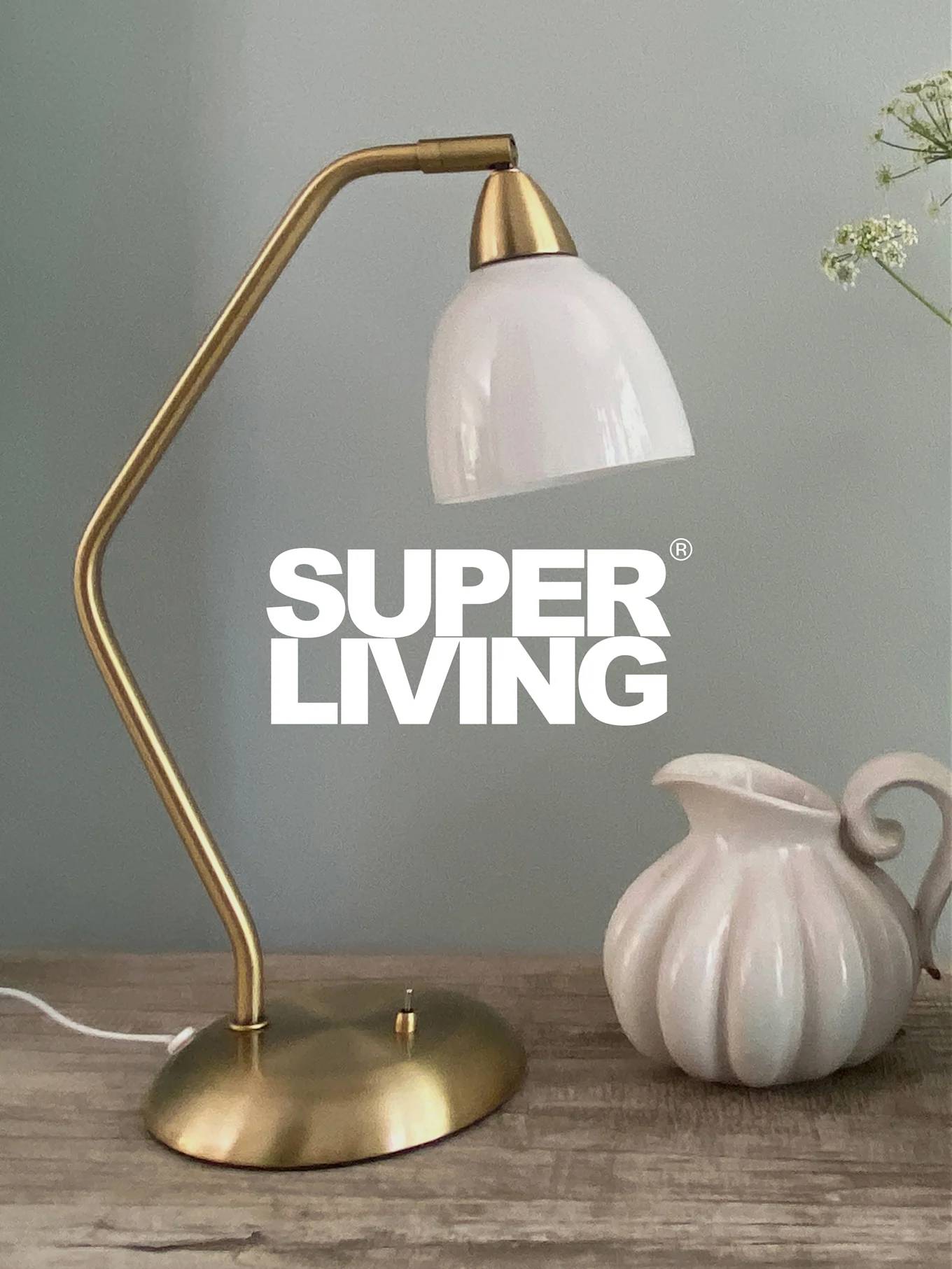 SUPER LIVING Branding über einer Messingtischlampe mit weißem Schirm und einem weißen, geriffelten Krug auf einem Holztisch.