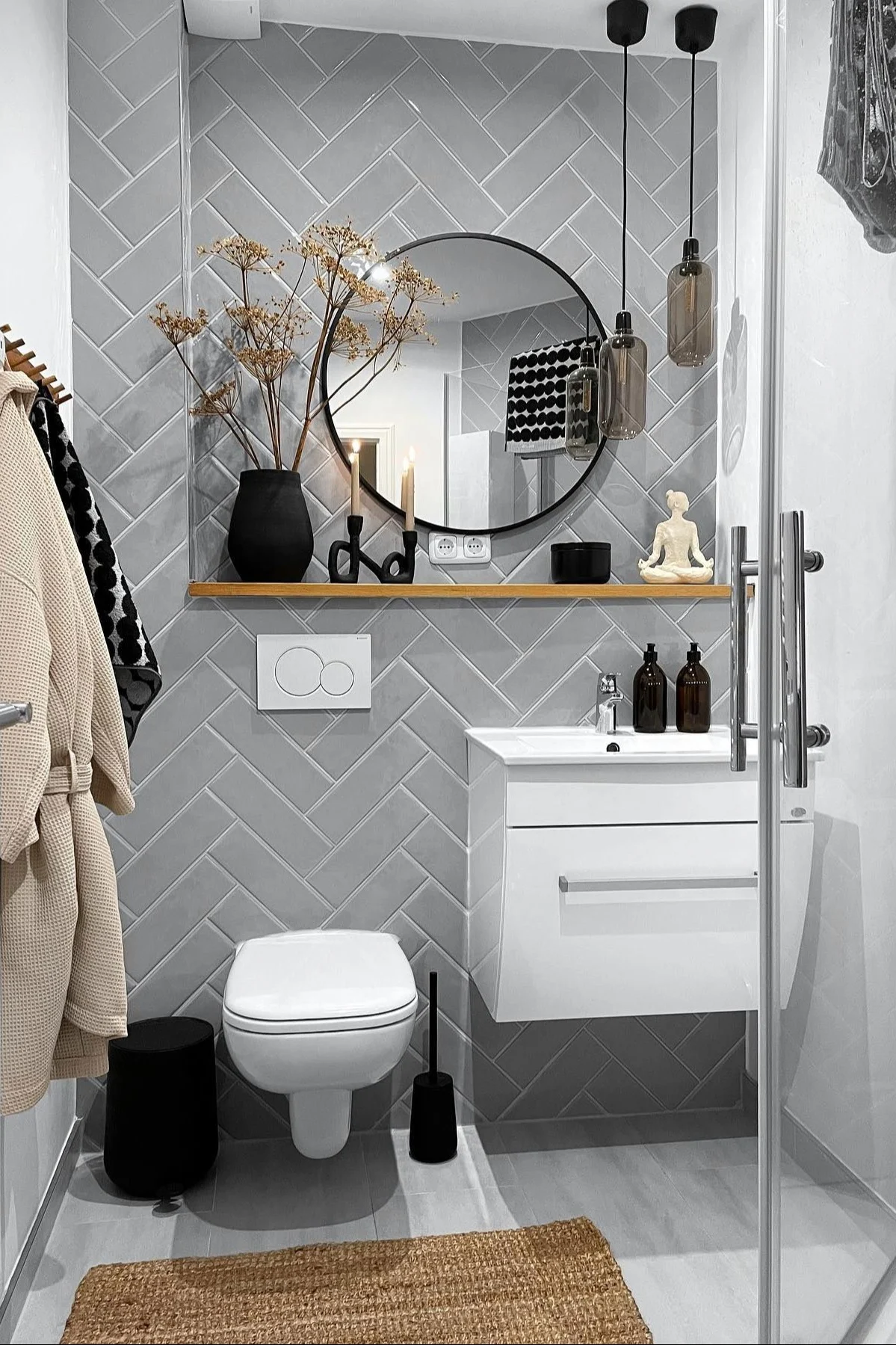 Modernes Badezimmer mit hellgrauen Fischgrät-Wandfliesen, einer weißen Toilette, Waschtisch, rundem Spiegel und einem Holzregal mit Deko.